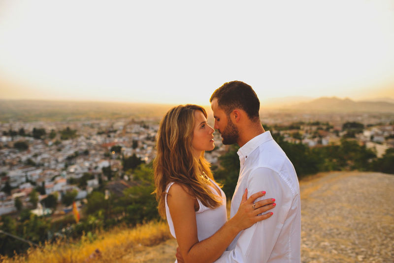 Side view of romantic couple standing on cobbled road against sky during sunset