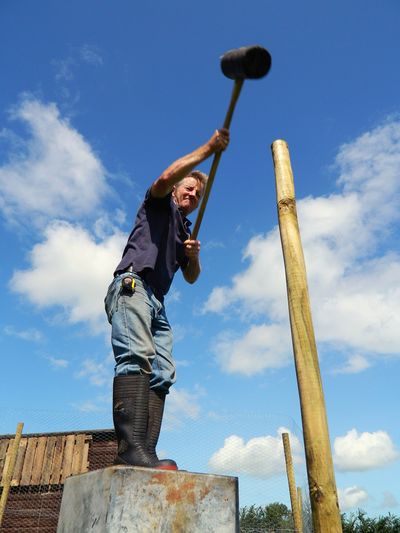 Low angle view of mature farmer hammering wooden post while standing against sky during sunny day
