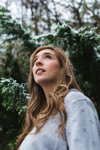 Low angle view of young woman looking away while standing by tree during snowfall