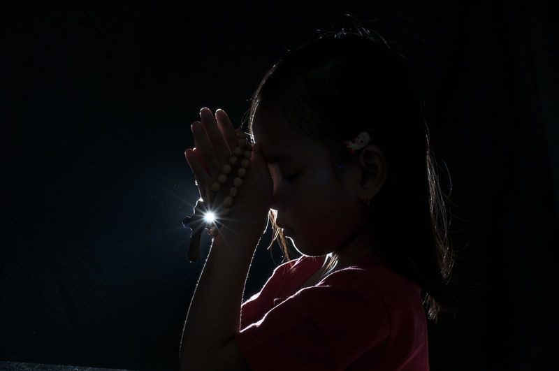 Cute girl with hands clasped praying against illuminated light