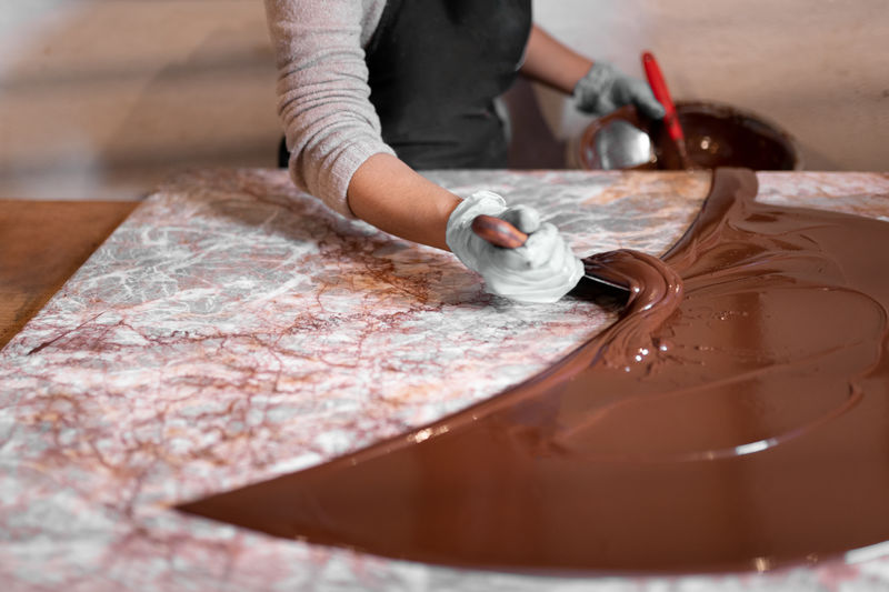 Anonymous employee in gloves using metal spatula to spread liquid chocolate on table while making dessert in confectionery