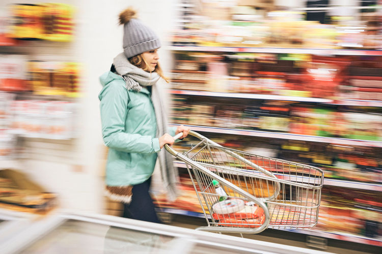 Side view of young female in warm clothes with shopping cart walking in supermarket and choosing food