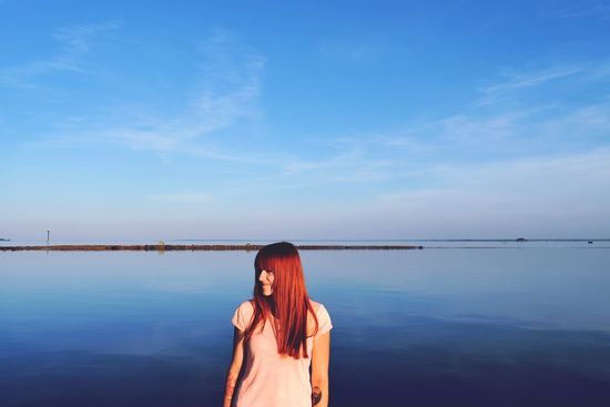 Redhead woman standing at beach against blue sky