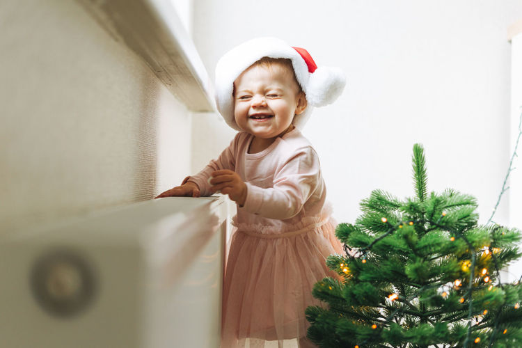 Cute little baby girl in pink dress and santa hat in room with christmas tree, happy new year