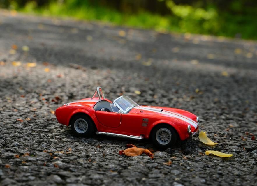 transportation, red, mode of transport, street, selective focus, land vehicle, car, focus on foreground, close-up, outdoors, road, toy, day, abandoned, high angle view, asphalt, no people, sunlight, surface level, ground