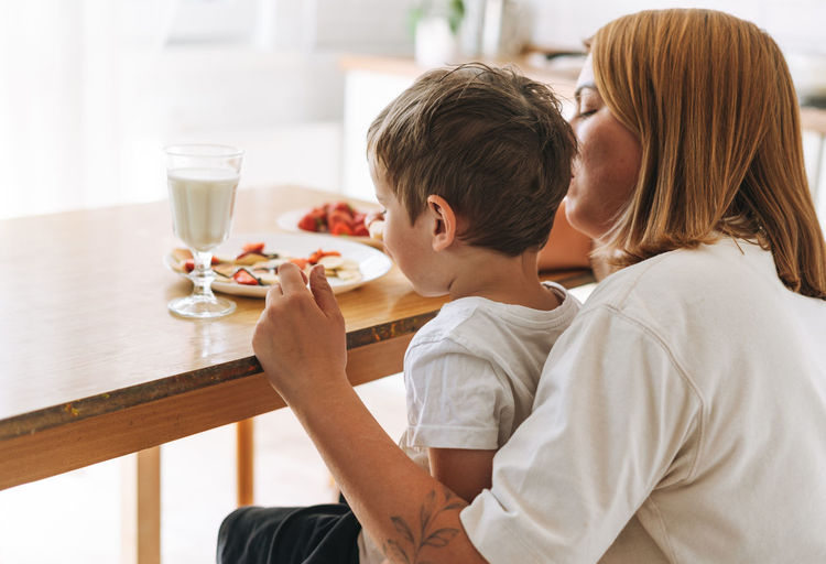 Cute toddler boy with his mother having breakfast with puncakes and berries in bright kitchen 