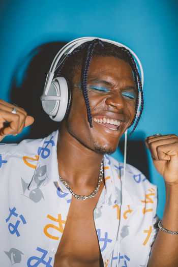 Optimistic african american male with bright makeup listening to music in headphones with closed eyes on blue background in studio