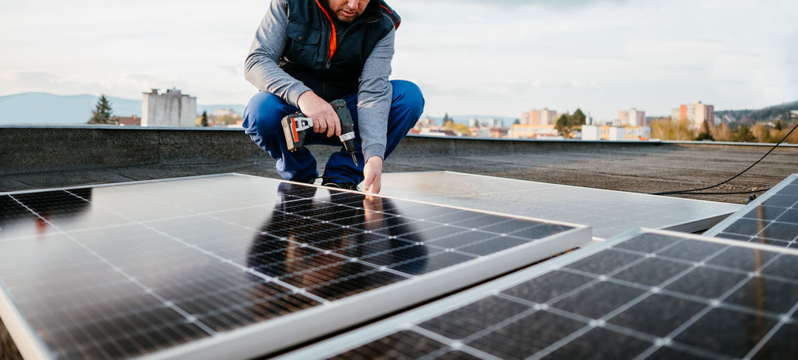 Man fixing solar panel through drill while crouching on rooftop