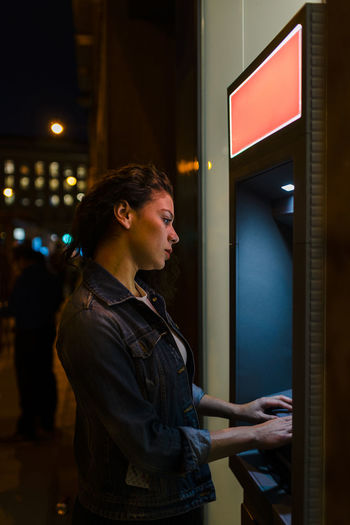 Young woman wearing denim jacket withdrawing money from atm in city at night