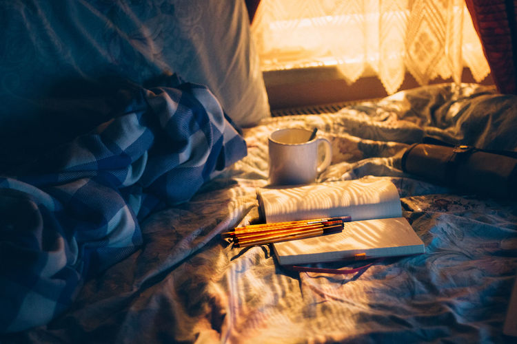 Coffee cup by book and pencils on bed by at home