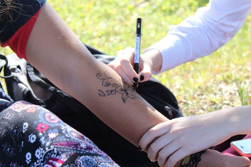 Cropped hands of friend drawing flowers on friends forearm sitting on grassy field