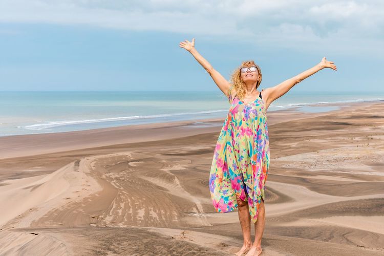 Mature woman with sunglasses in colorful dress raising the arms at the top of sand dune