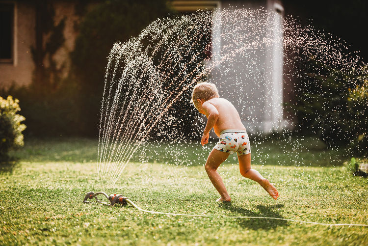 Young white boy running under the water from the sprinkler in garden
