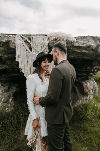 Delighted newlywed couple in stylish clothes standing against macrame hanging on rock in nature on wedding day and embracing
