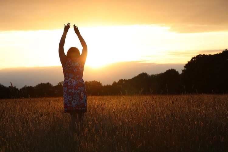 Woman with arms raised on field against sky during sunset