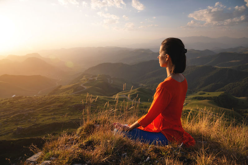 Young caucasian woman meditates in the lotus position in the grass in the mountains near the cliff