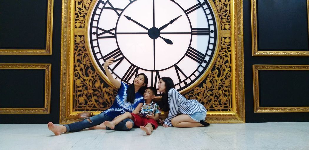Full length of woman taking selfie with family while sitting on tiled floor against clock