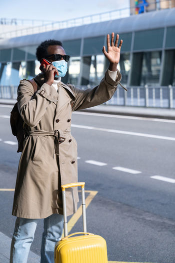 Afro traveler man wear face mask stands in airport terminal, talk on phone, calling taxi. covid-19