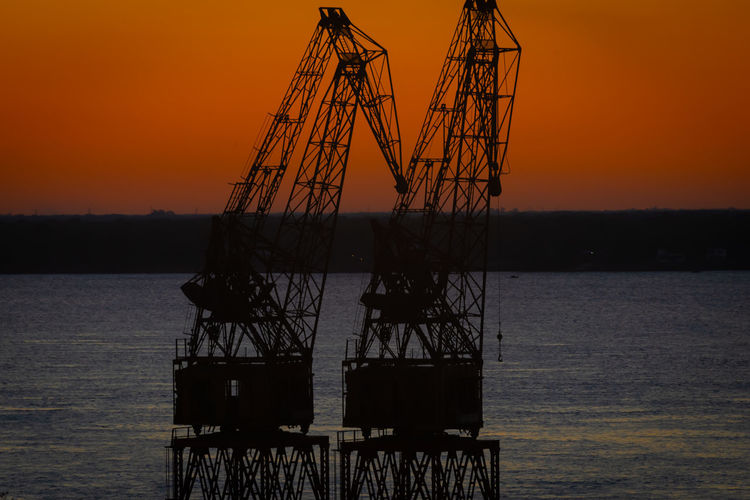 Silhouette cranes on pier by sea against sky during sunset