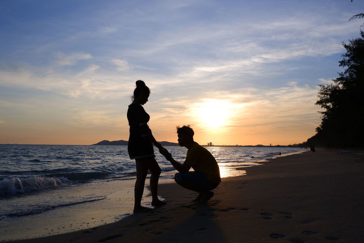 Silhouette boyfriend proposing girlfriend at beach against sky during sunset