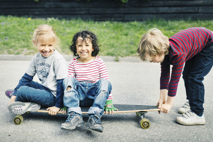 Portrait of happy children sitting on skateboard while friend pulling it at yard