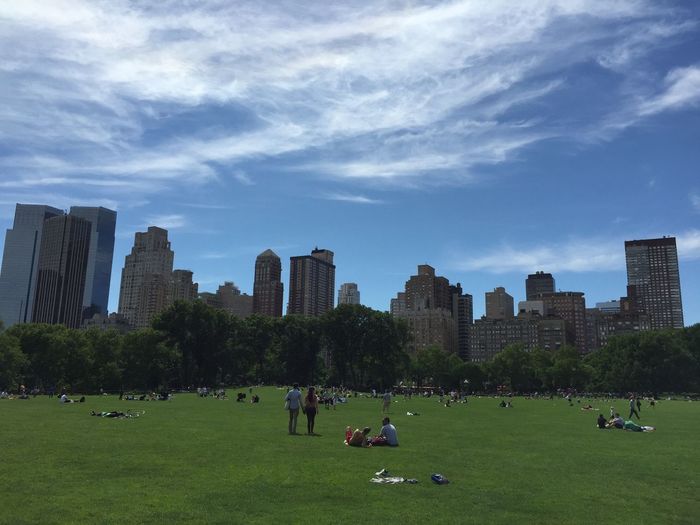 People enjoying on grassy field at park by cityscape against sky