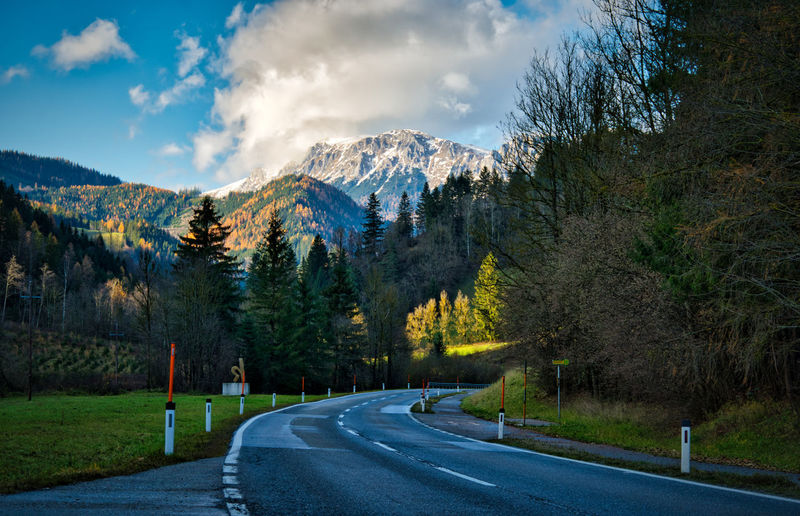 Empty road by trees and mountains against sky