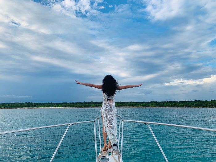 Rear view of woman with arms outstretched standing on boat in sea against sky