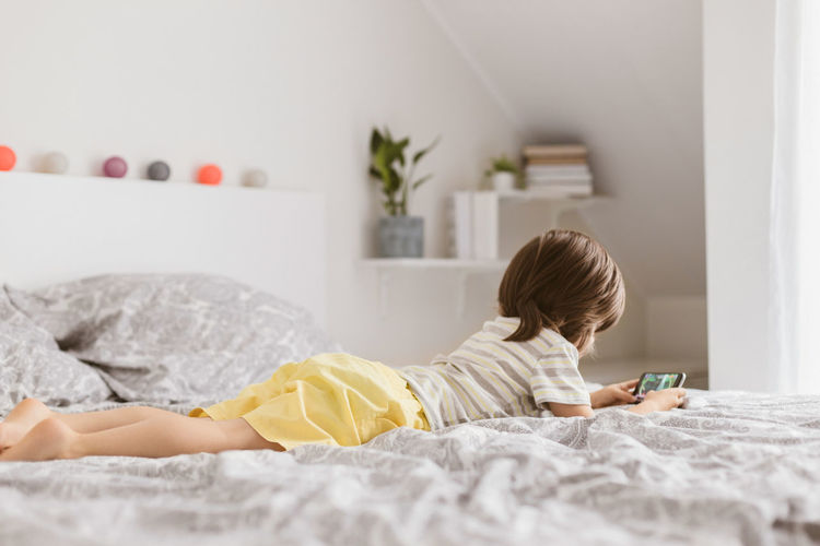 A cute boy, toddler, lies on the bed in the bedroom and plays with the phone, smartphone.