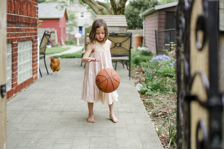 A small girl dribbles basketball in backyard with a chicken behind her