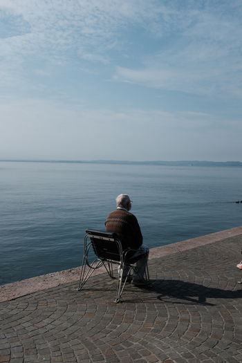 Rear view of man sitting on bench by sea against sky