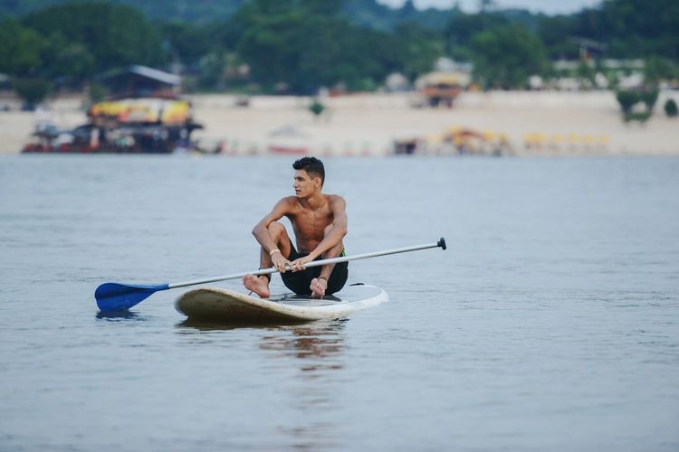 Full length of shirtless man paddleboarding on sea