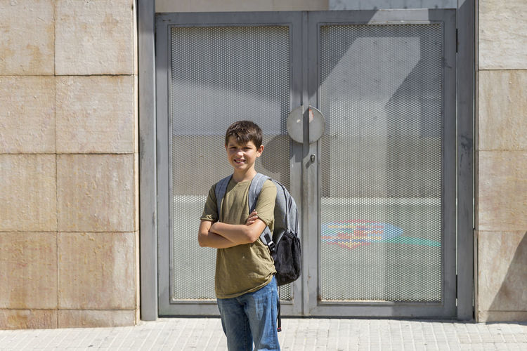 Portrait of smiling boy with backpack standing against metal door during sunny day