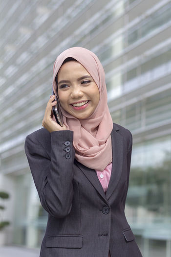 Happy businesswoman using smart phone while standing against office building