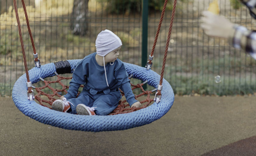 Cute little boy with down syndrome in a funny hat walks in the playground, swinging on a swing