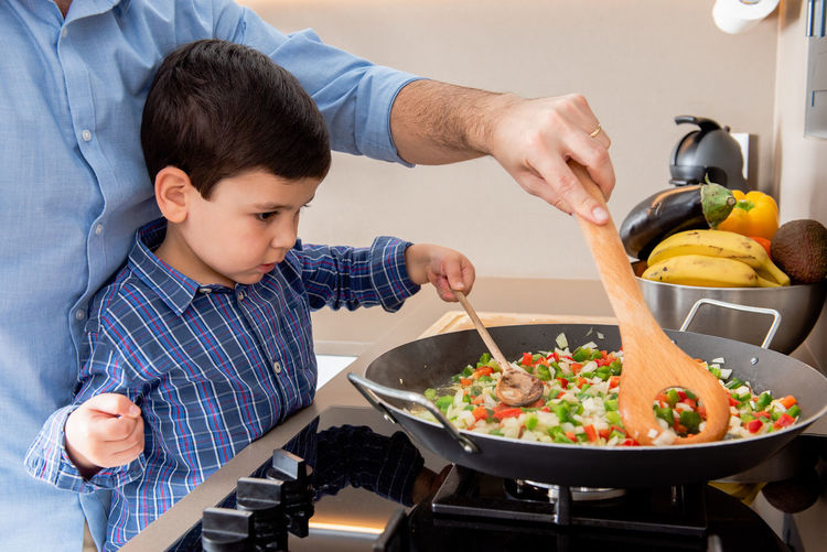 Happy young father and cute son sauteing cut ripe vegetables in frying pan while cooking lunch together in contemporary kitchen