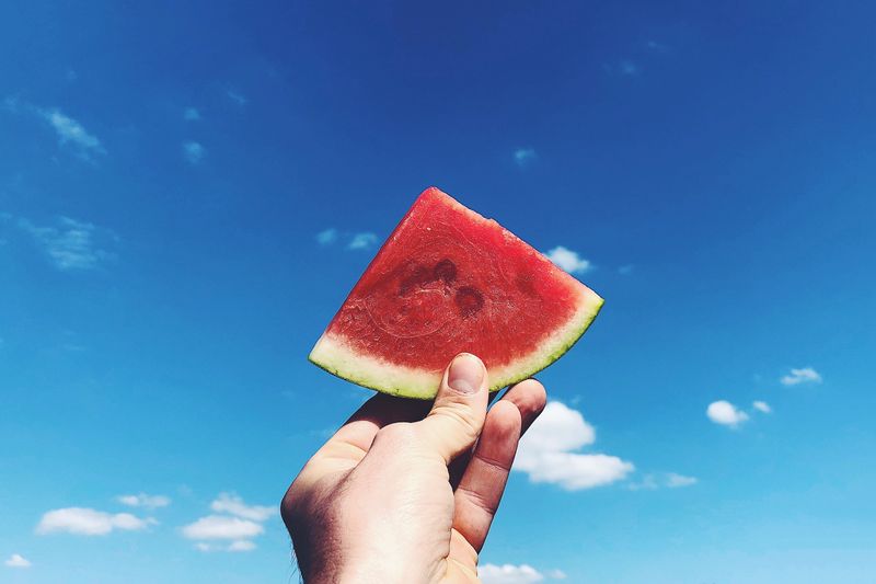 Cropped hand holding watermelon slice against blue sky