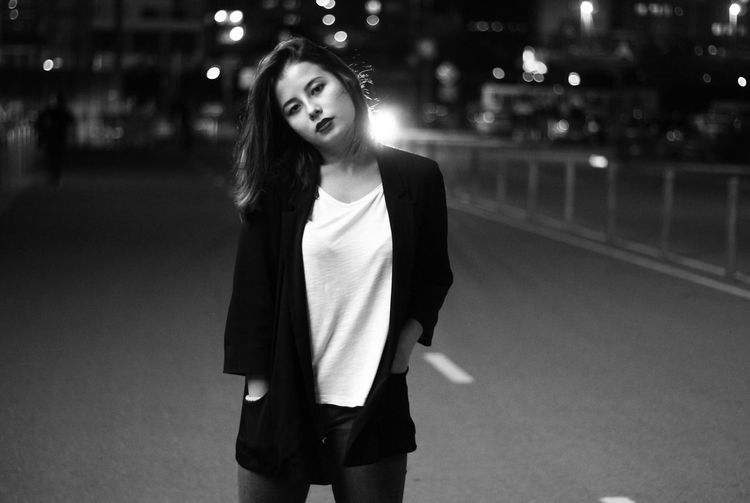 Confident young woman standing with hands in pockets on city street at night