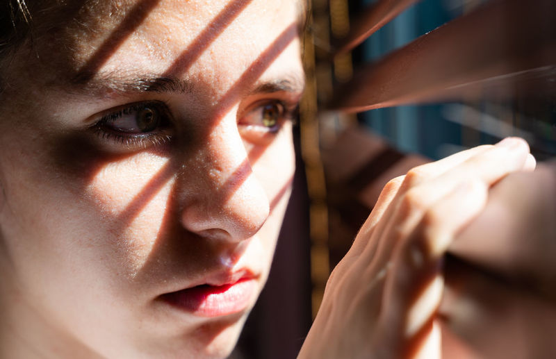 Youth girl with green eyes opening a wooden curtain with her fingers to look through the window while the sunlight creates sun and shadow on her face that shows hopeless. horizontal photo