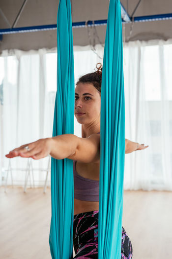 Active female in sportswear practicing warrior pose on hanging hammock during aerial yoga training in light spacious studio with windows