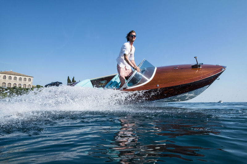 Low angle view of young man riding speedboat on sea against clear blue sky