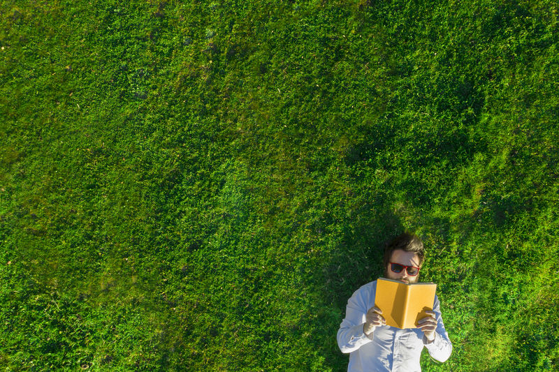 Bearded man with sun glasses lying on the grass reads a yellow book. aerial photo taken from a drone