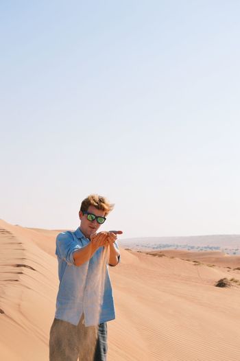 Young man falling sand while standing on desert against clear sky during sunny day