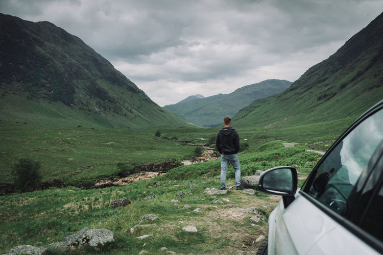 Rear view of man by car on field amidst mountains against cloudy sky