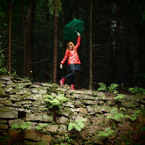 Happy woman in raincoat with umbrella standing on stone wall at forest