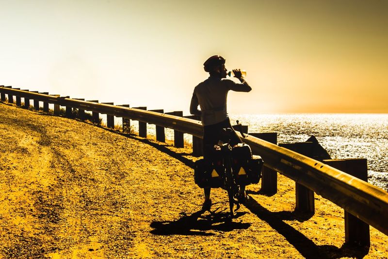 Man on bicycle standing by railing against sky during sunset