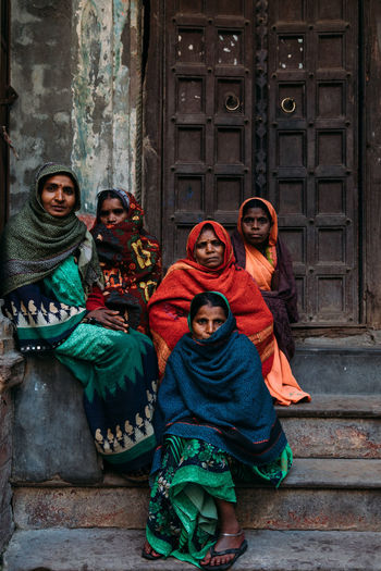 Varanasi, india - february, 2018: group of serious hindu women in colorful traditional clothes with scarfs on heads sitting or standing together next to door of flaky building