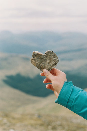 Cropped hand holding heart shape stone against mountains