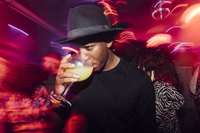 Low angle of african american guy sipping alcohol cocktail under colorful illumination during party in nightclub
