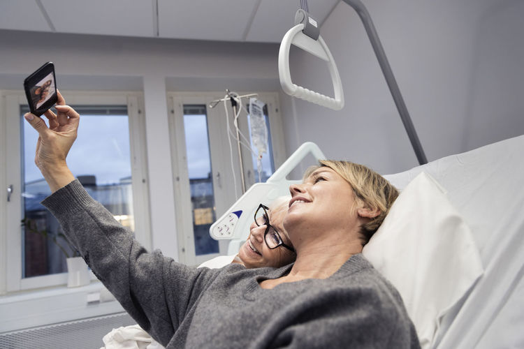 Adult daughter taking selfie with mother on hospital bed